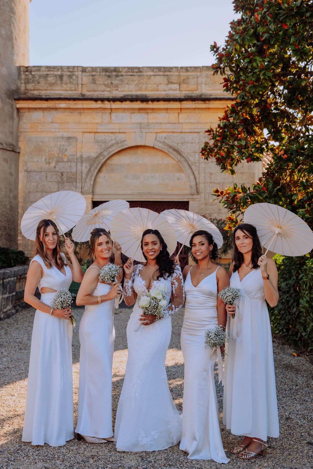 La mariée et les demoiselles d'honneur prennent la pose pour les photos de groupes. Les filles sont en blanc et chacune a une petite ombrelle